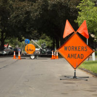 Orange and Black "Workers Ahead" Sign in Residential Neighborhood with Equipment in Background, Daytime Orange and Black "Workers Ahead" Sign in Residential Neighborhood with Equipment in Background, Daytime