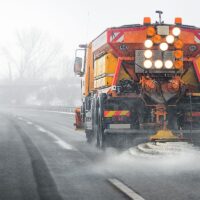 Snow plow salting street in winter time. Orange truck deicing. Maintenance winter vehicle back side.