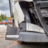 Close-up of a damaged truck front on a roadside, with a tow truck and traffic sign visible. Overcast sky and trees in the background.
