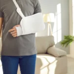 A person in a gray shirt and blue jeans stands with their right arm in a white sling, suggesting injury. They are in a bright, modern living room.